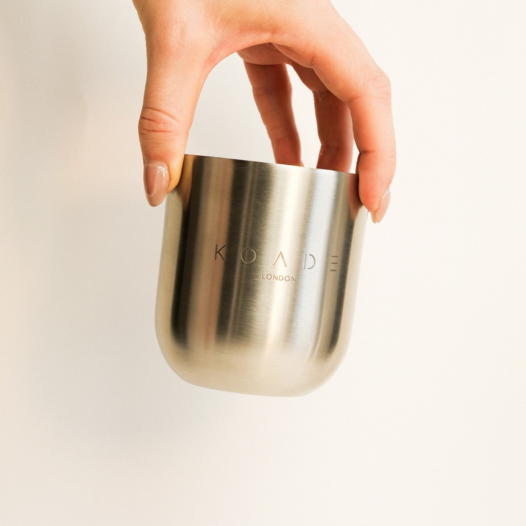 Hand holding a metallic cup with 'Koade London' branding on a light background