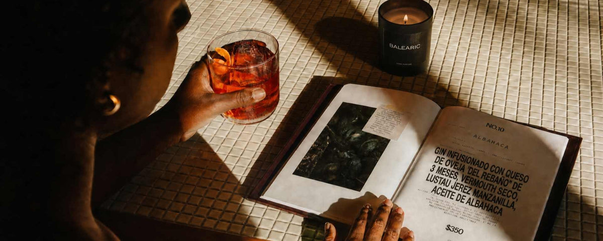A person seated at a table holds a glass of amber-colored drink beside an open book with printed text and a candle. Strong sunlight casts deep shadows across the table and book pages.