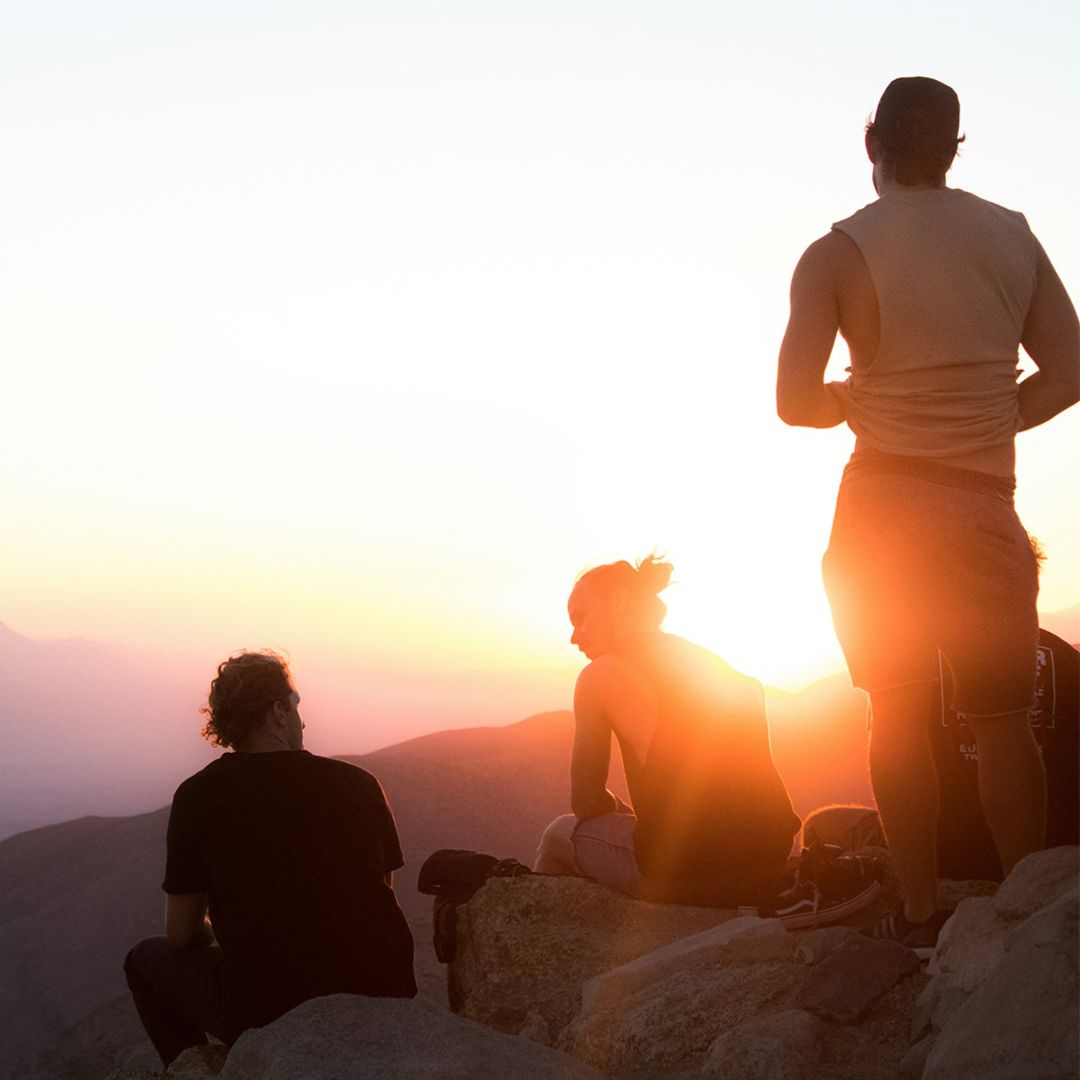 Three people sitting on a mountain top at sunset