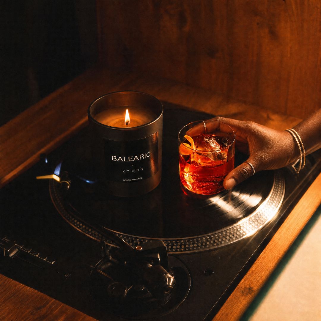 Candle labeled 'BALEARIC' next to a glass of orange liquid on a dark surface.