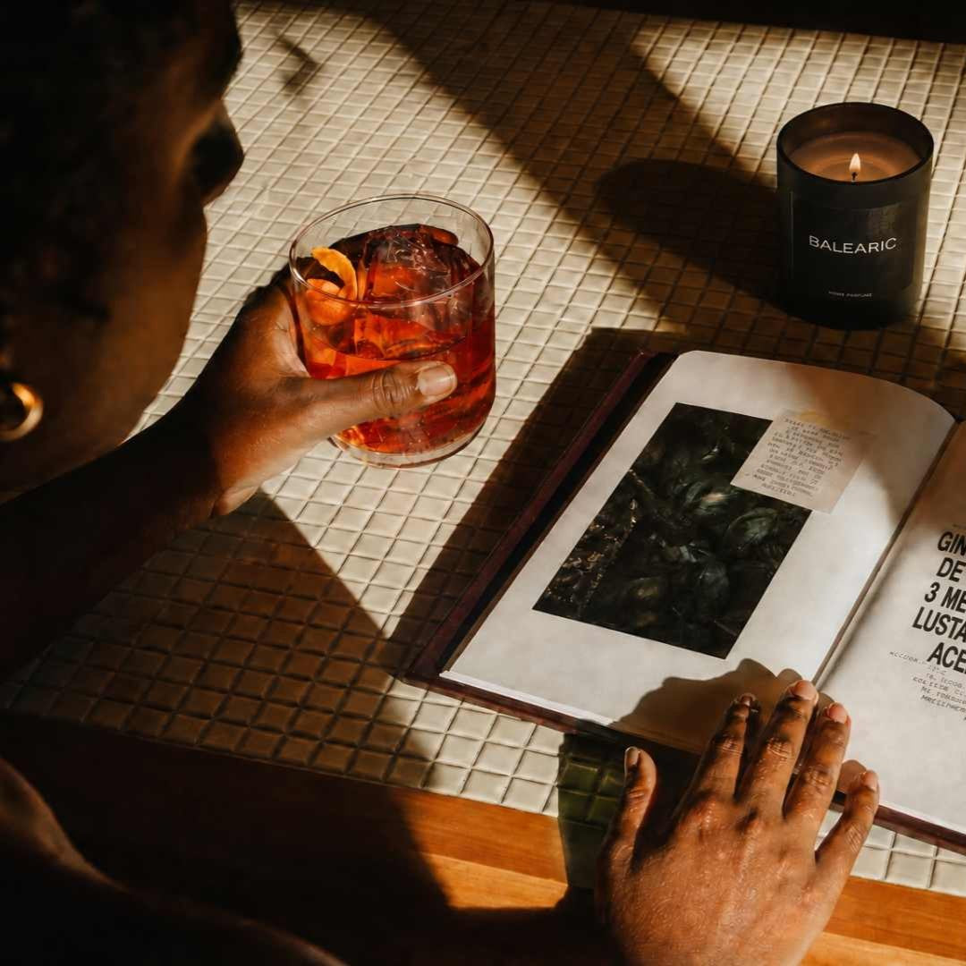 A person seated at a table holds a glass of amber-colored drink beside an open book with printed text and a candle. Strong sunlight casts deep shadows across the table and book pages.