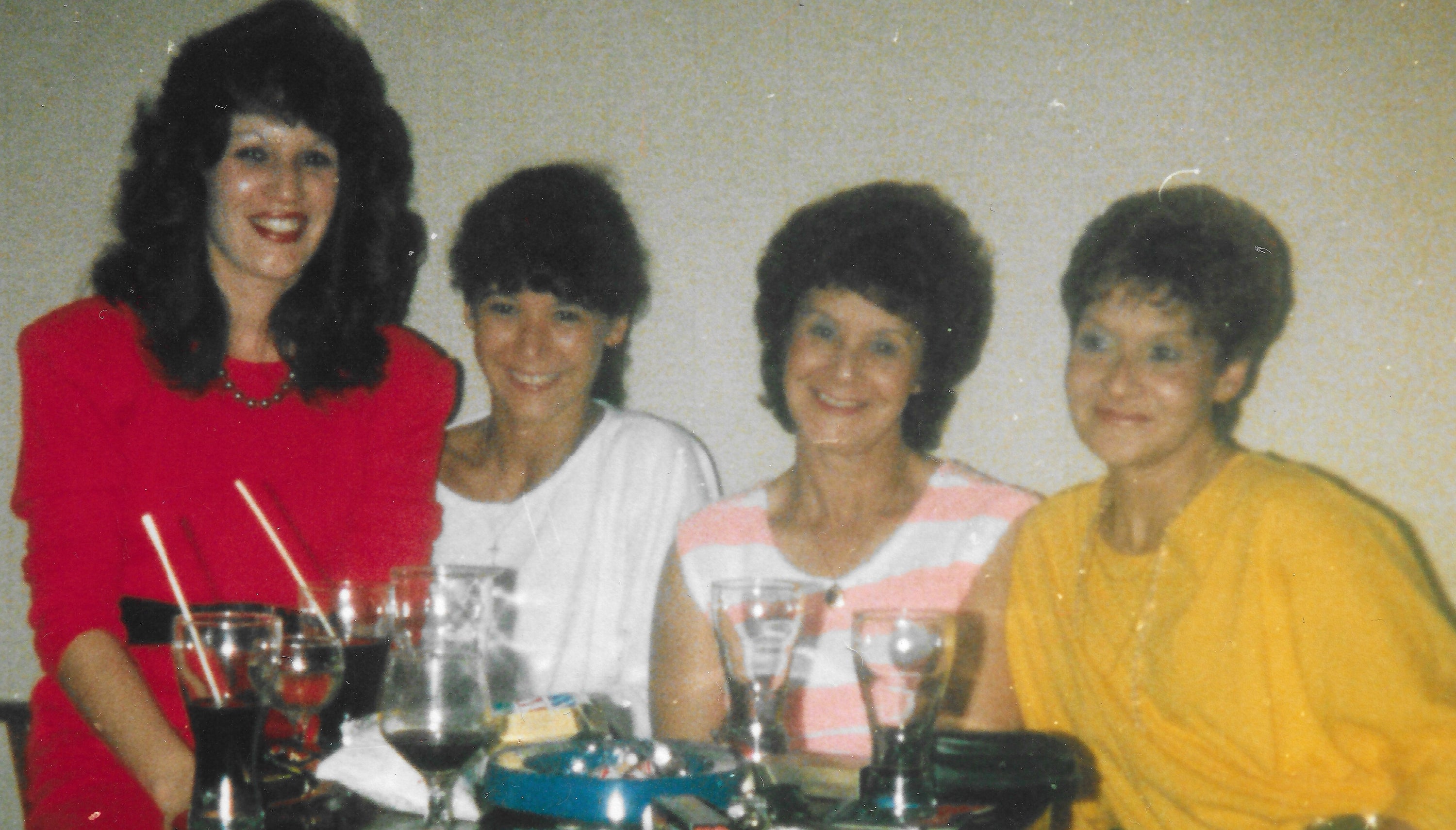 Four women sitting together at a table with drinks and food, smiling for the camera.