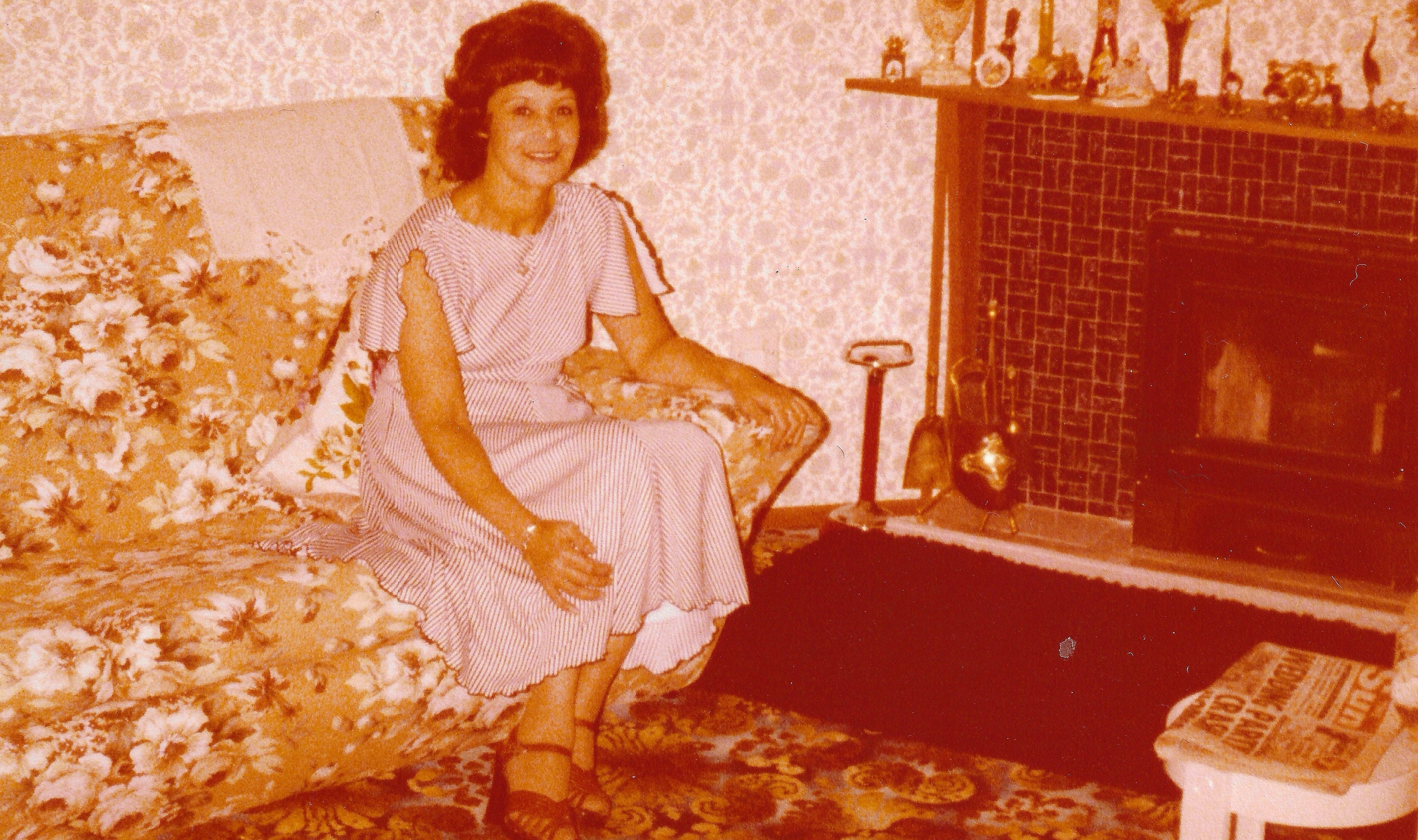 Woman in a vintage dress sitting on a floral-patterned couch in a room with a fireplace.