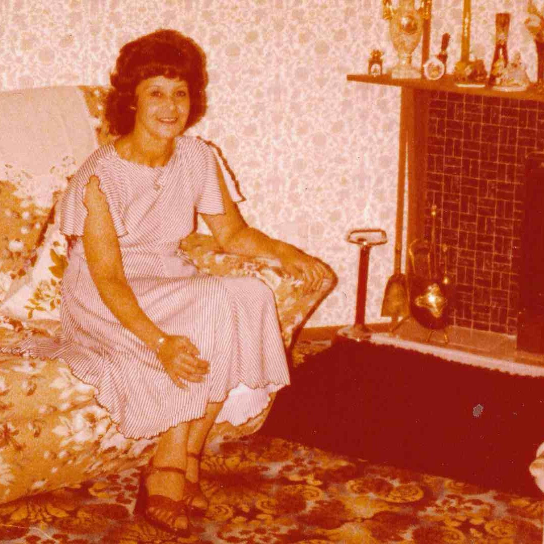 Woman sitting on a floral-patterned chair in a room with a fireplace and decorative items.
