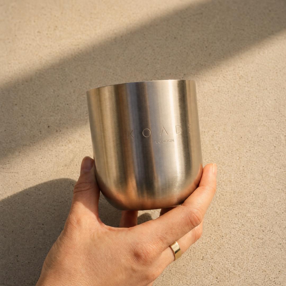 Hand holding a silver cup with 'KOADE London' branding on a sandy background