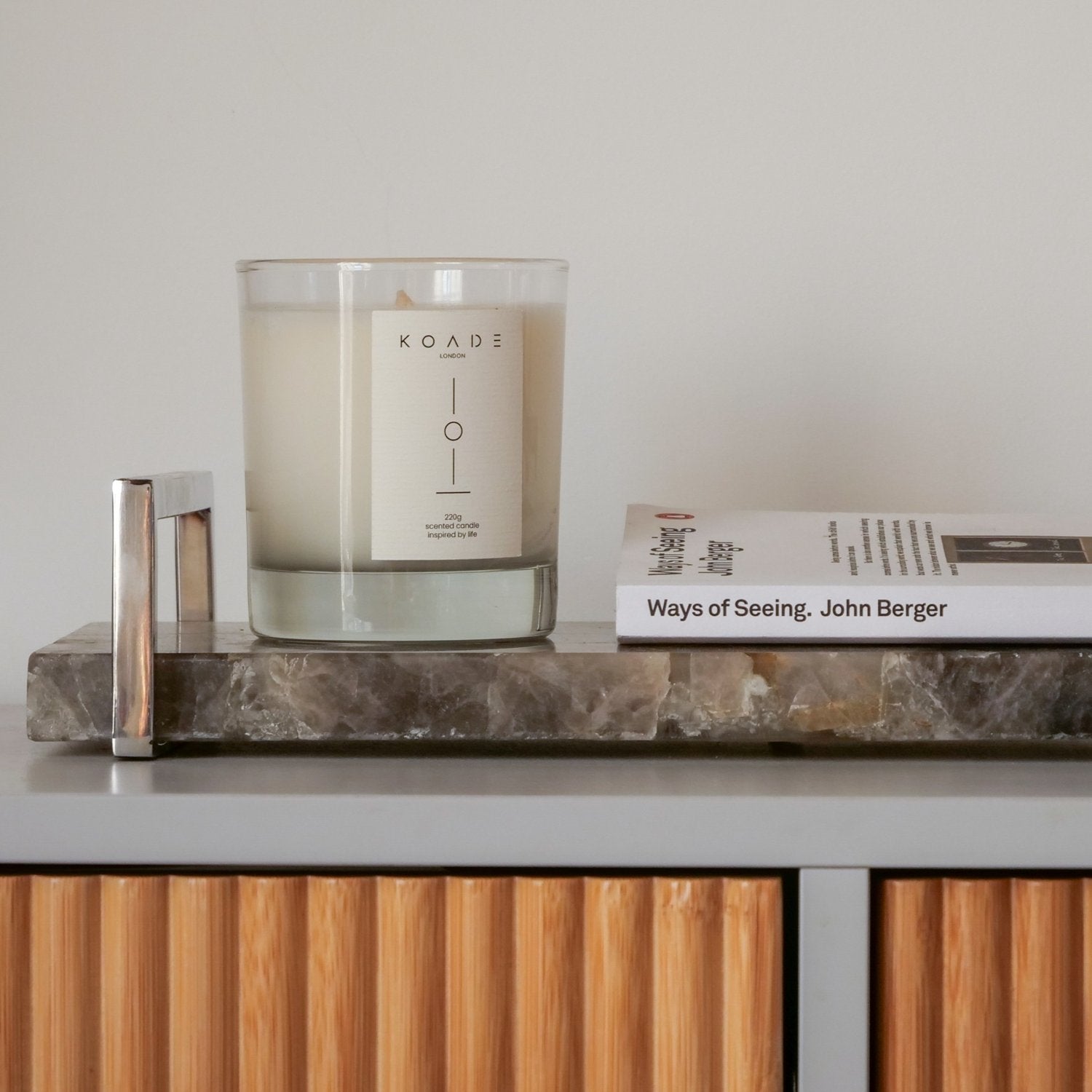 Candle with a KOADE label on a marble tray next to a book on a wooden shelf.