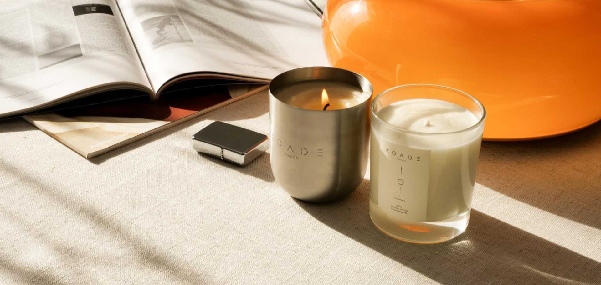 Two candles on a table with an open book and orange object in the background