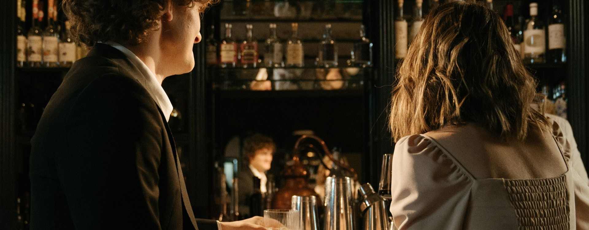 A dimly lit bar scene with two people facing each other behind the counter, with shelves of bottles in the background. The person on the left wears a dark suit, and the person on the right wears a light-colored blouse.