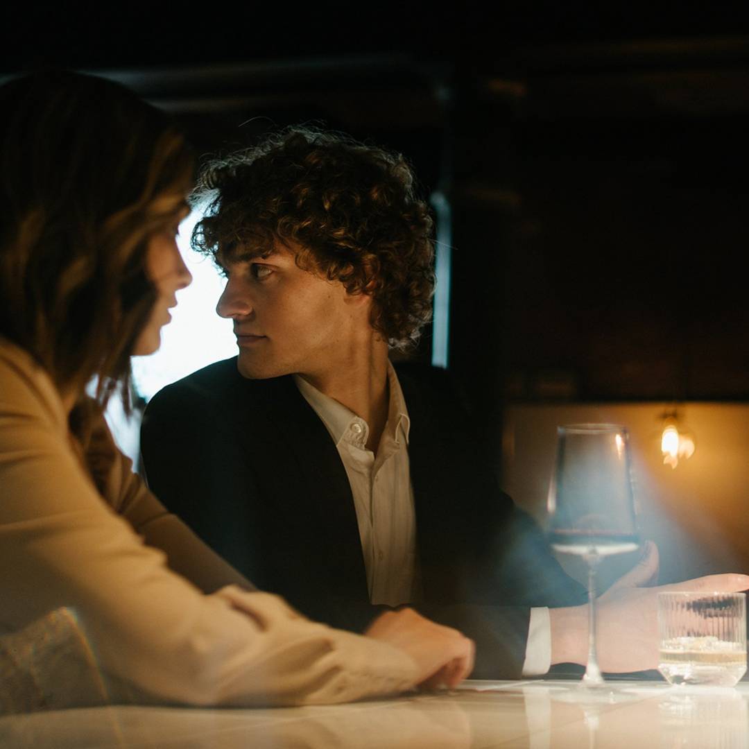 Two people sitting close together in a dimly lit room with a glass of wine on the table.
