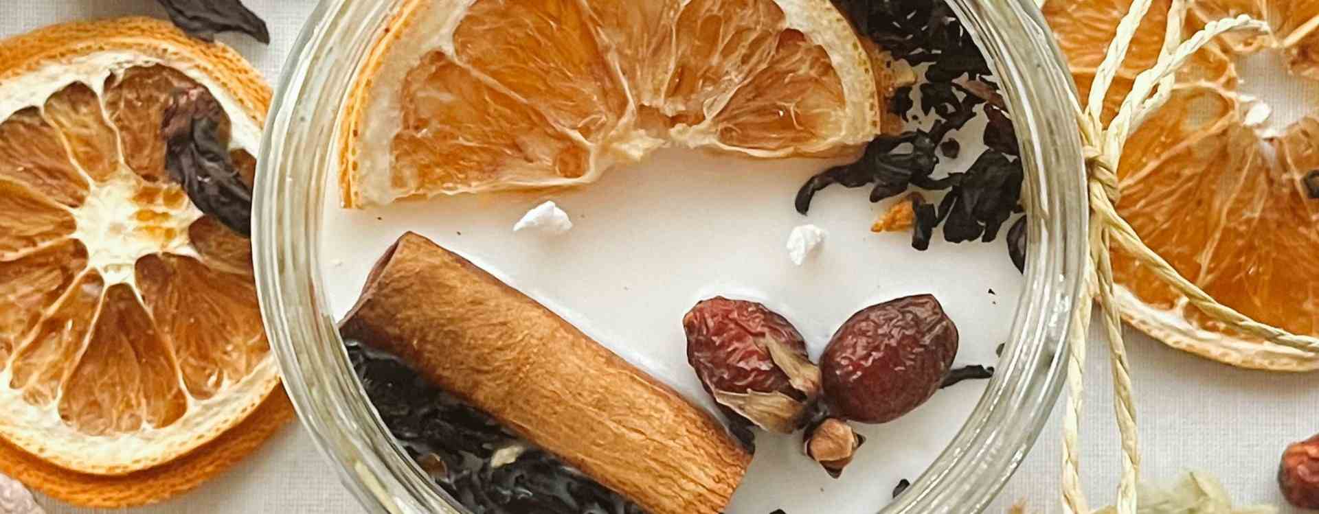 A clear glass jar filled with a warm herbal tea blend, featuring dried orange slices, a cinnamon stick, dark berries, and small dried flowers, surrounded by loose dried orange slices on a light surface.