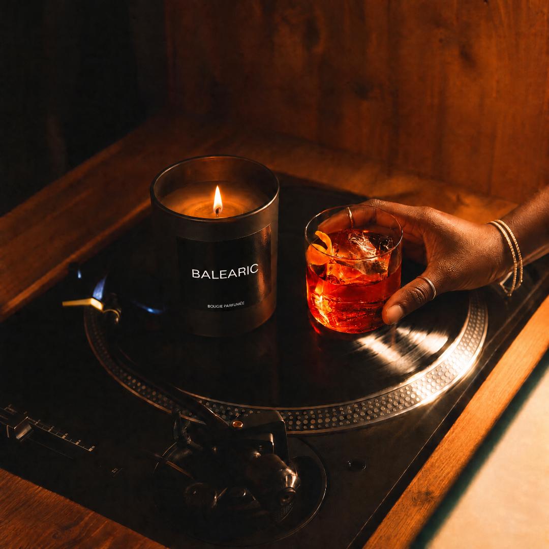 A dark metal candle jar labeled BALEARIC sits on a wooden record player next to a hand holding an amber cocktail with a citrus twist. Warm ambient lighting casts a blue glow on the turntable and background wood.