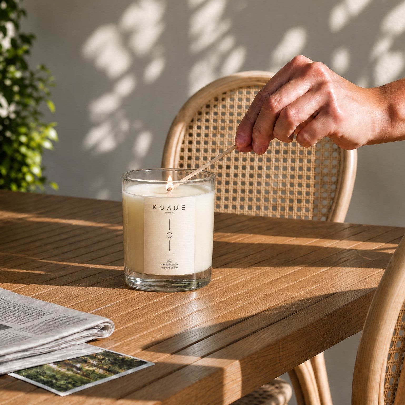 Hand lighting a candle labeled 'Koade' on a wooden table with a wicker chair and plant in the background.