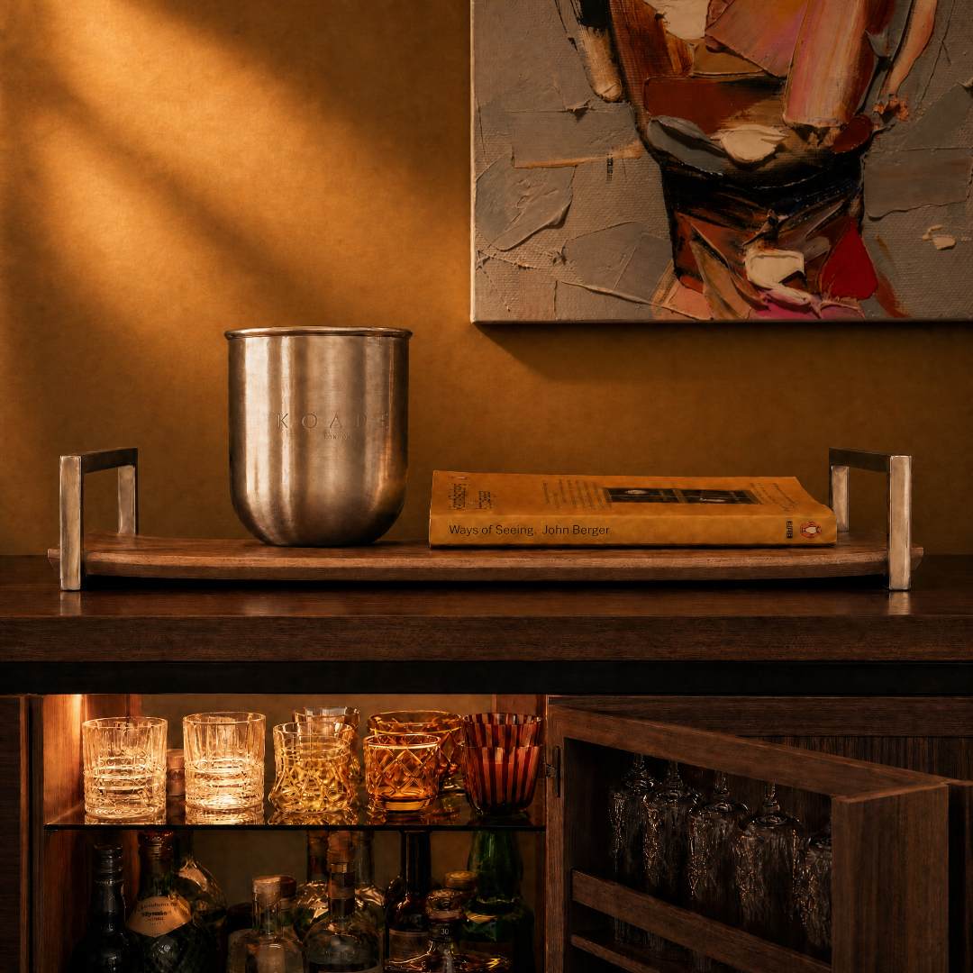 Metal candle holder on a wooden tray with a book and abstract painting in a warm-toned room.