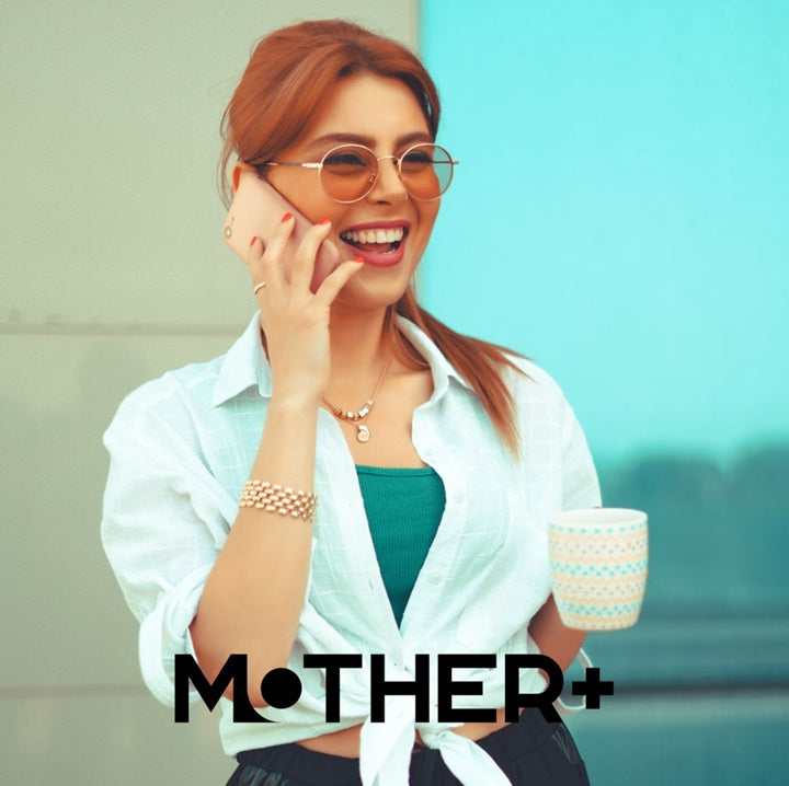 Woman with sunglasses holding a cup, standing against a light blue wall with 'MOTHER+' branding.