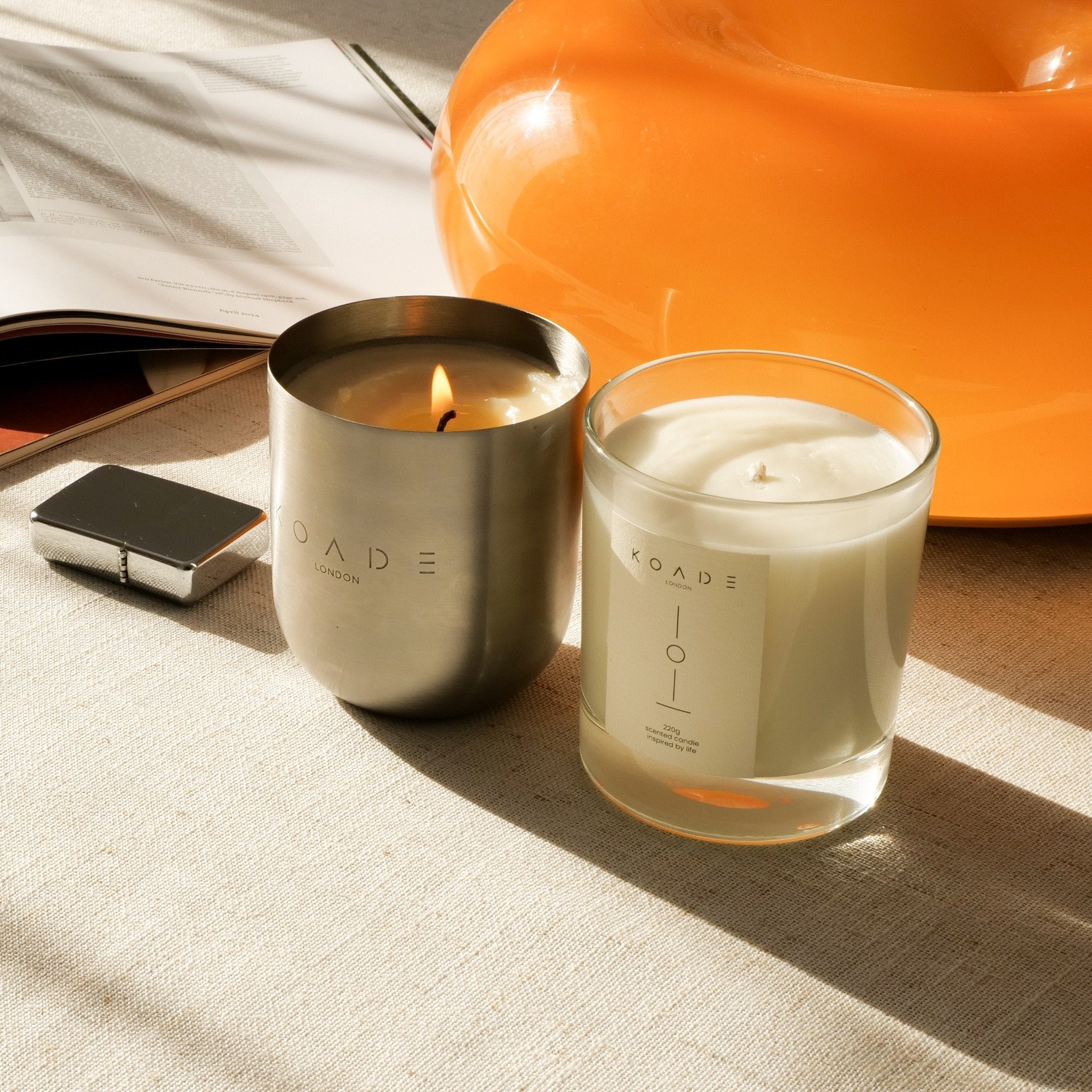 Two candles, one in a metallic container and the other in a glass jar, on a textured surface with an orange object in the background.